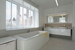 White bathroom with white window coverings over large sunny windows.