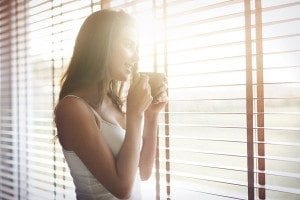 Woman standing by a window with a cup of coffee looking out past her venetian blinds.